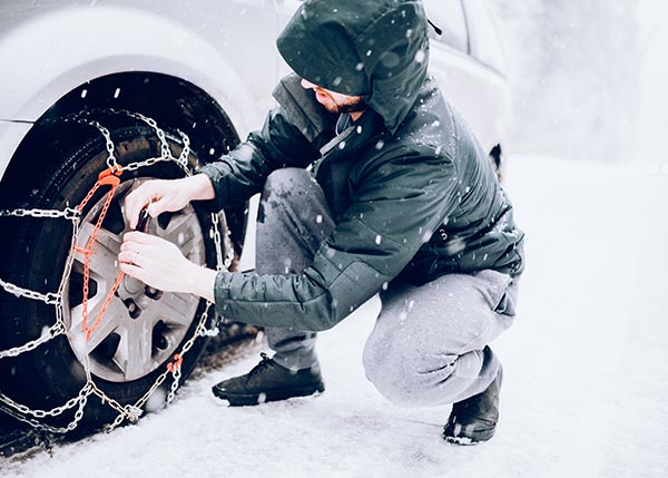 A low angle view of a front wheel drive vehicle on a snowy and ice covered road, a man putting chains on the wheels to improve traction in the winter weather conditions. Horizontal image with copy space.